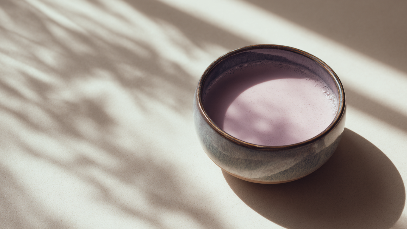 Bol en céramique rempli d’un latte à l’ube violet pâle, posé dans la lumière du matin avec des ombres végétales douces sur une surface beige. Une scène minimaliste et apaisante, évoquant un moment de bien-être. Image