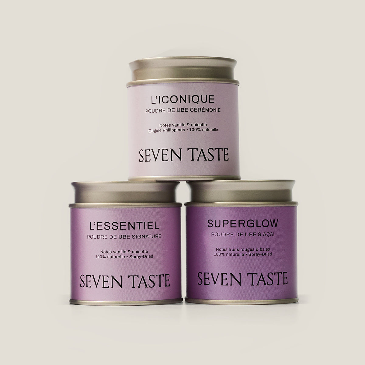 Trois boîtes de poudre d’ube Seven Taste empilées en pyramide, L’Iconique, L’Essentiel et Superglow, sur fond beige clair.